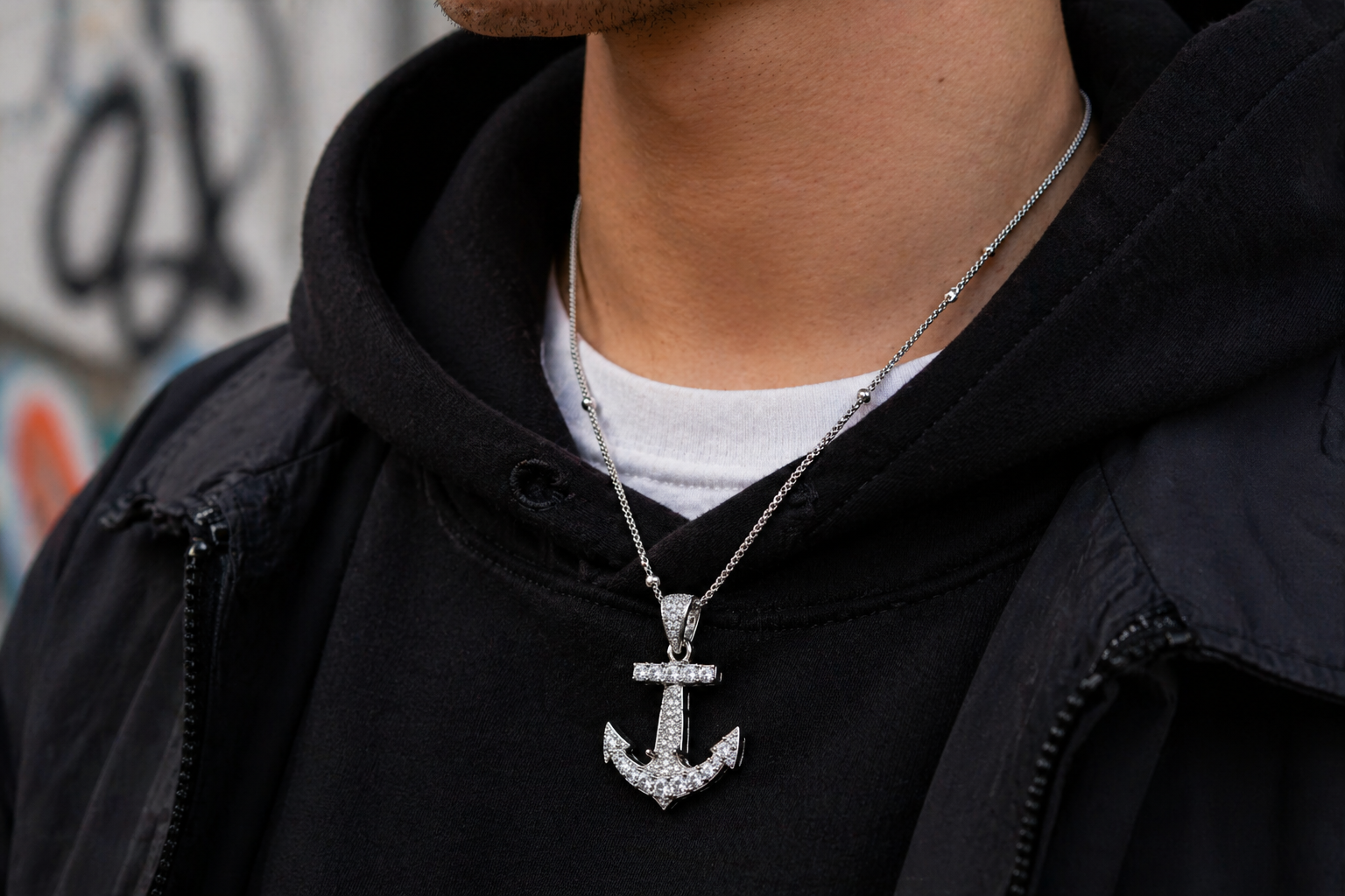 Person wearing a black hoodie with a silver anchor pendant necklace against a graffiti-covered wall.