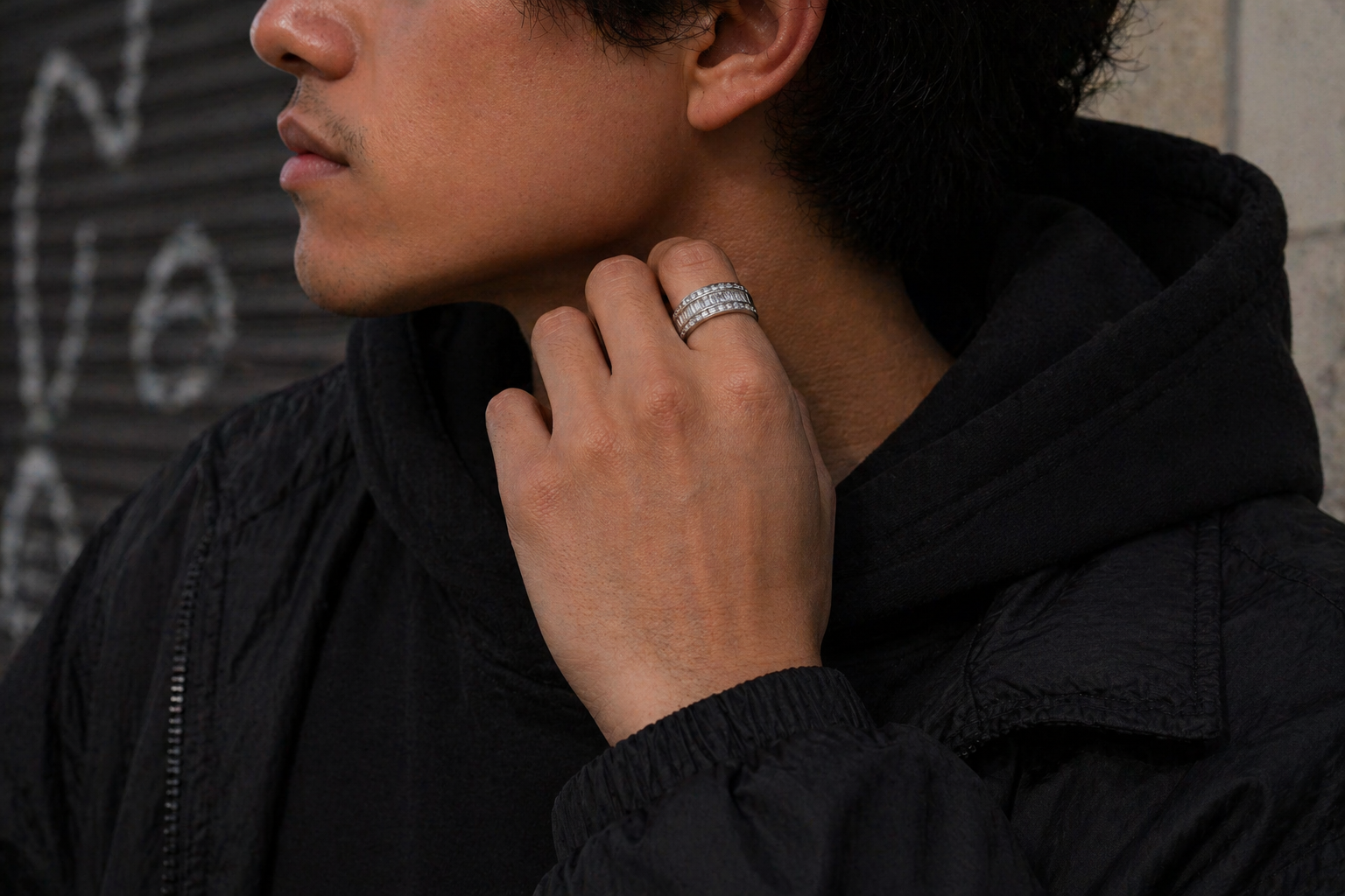 Man wearing a black jacket with a ring on his finger, standing against a dark background.